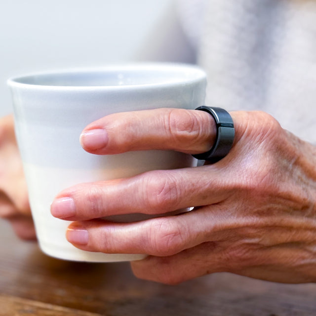 A hand wearing a graphite Lotus ring on the index finger holding a white mug.