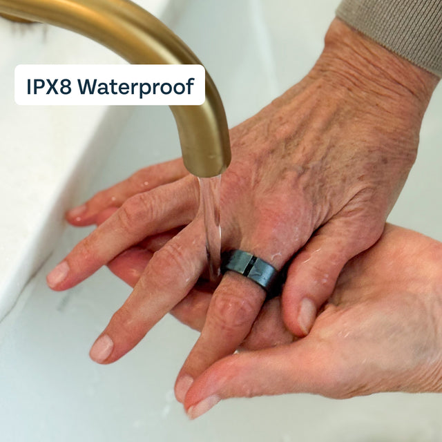 Hands rinsing with the graphite Lotus ring under running water from a gold faucet in a white sink. Label states IPX8 Waterproof.